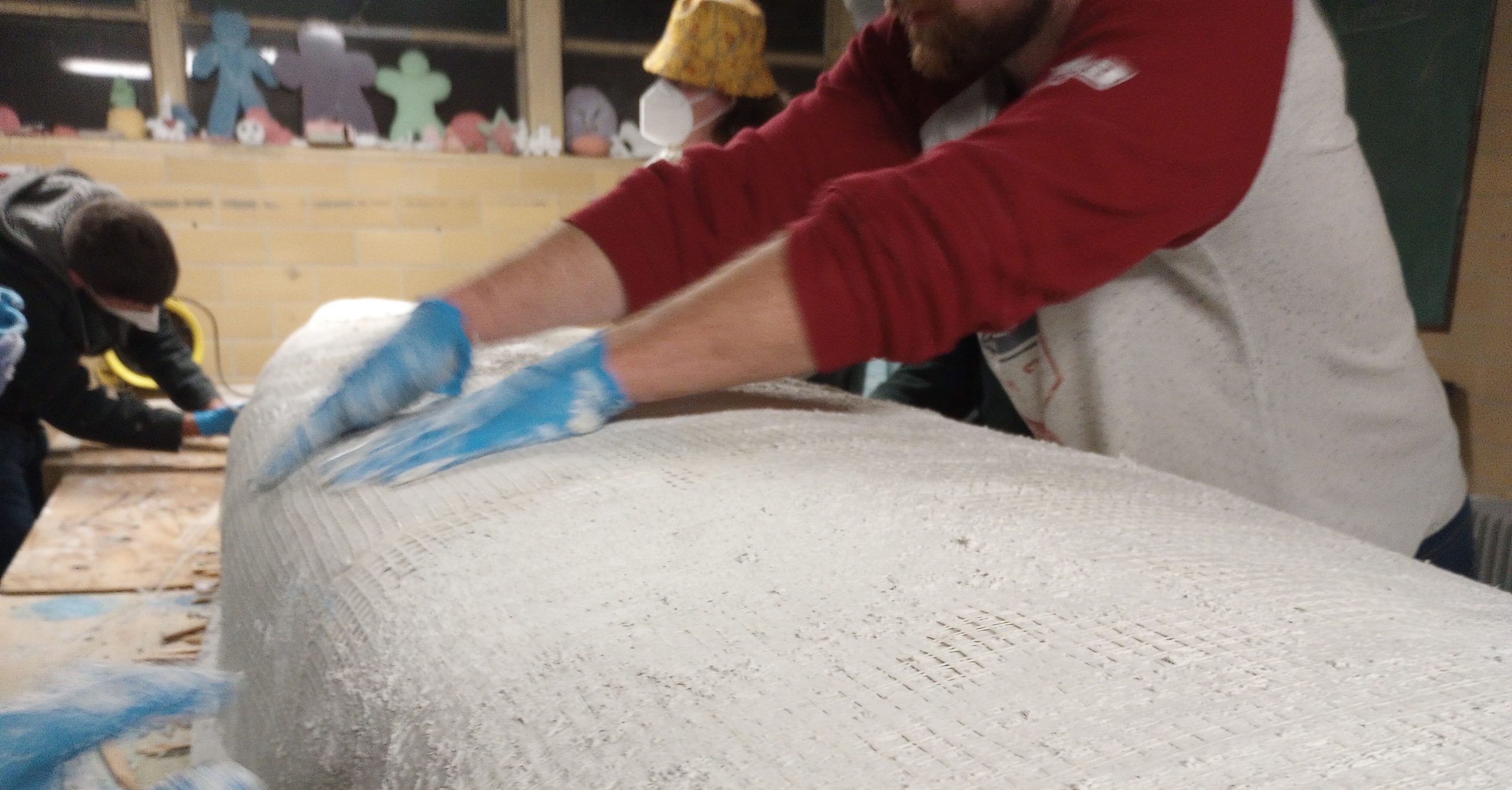 person in the process of adding fiber mesh on top of the first white layer of concrete on the canoe